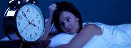 Woman lying awake in bed staring at an alarm clock at night, from SANUSq Health on the effects of chronic insomnia
